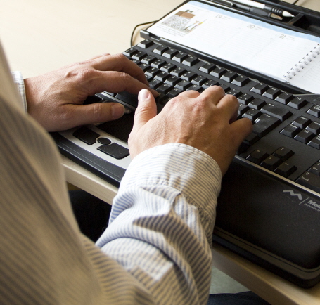 En närbild på ett par händer som skriver på ett svart tangentbord. Ovanför tangenterna ligger en anteckningsbok. I bakgrunden syns en skrivbordsskiva i trälaminat. Foto.