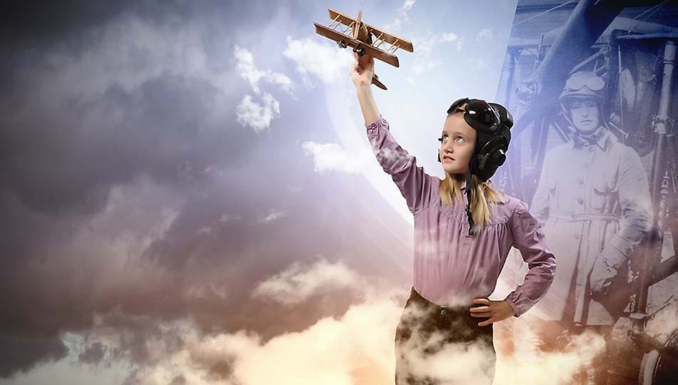 En flicka i flygkläder håller upp ett flygplan i trä med ena armen och kollar uppåt. Foto. 