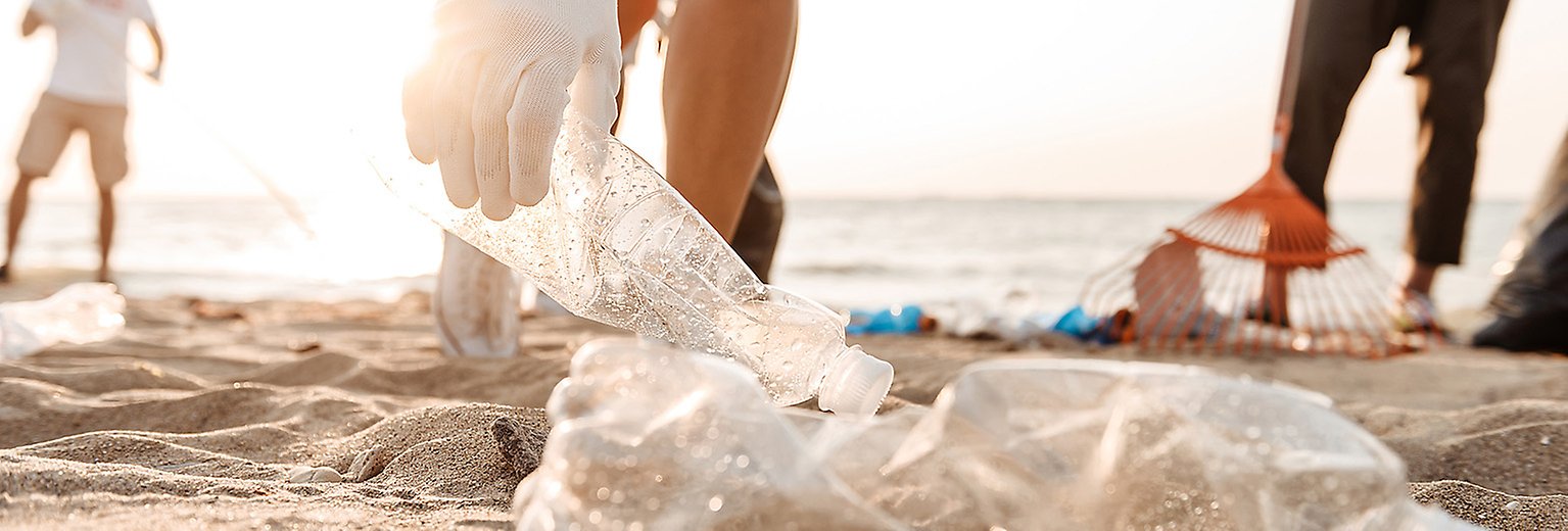 En person plockar upp en plastflaska på stranden tillsammans med två andra.  Foto. 