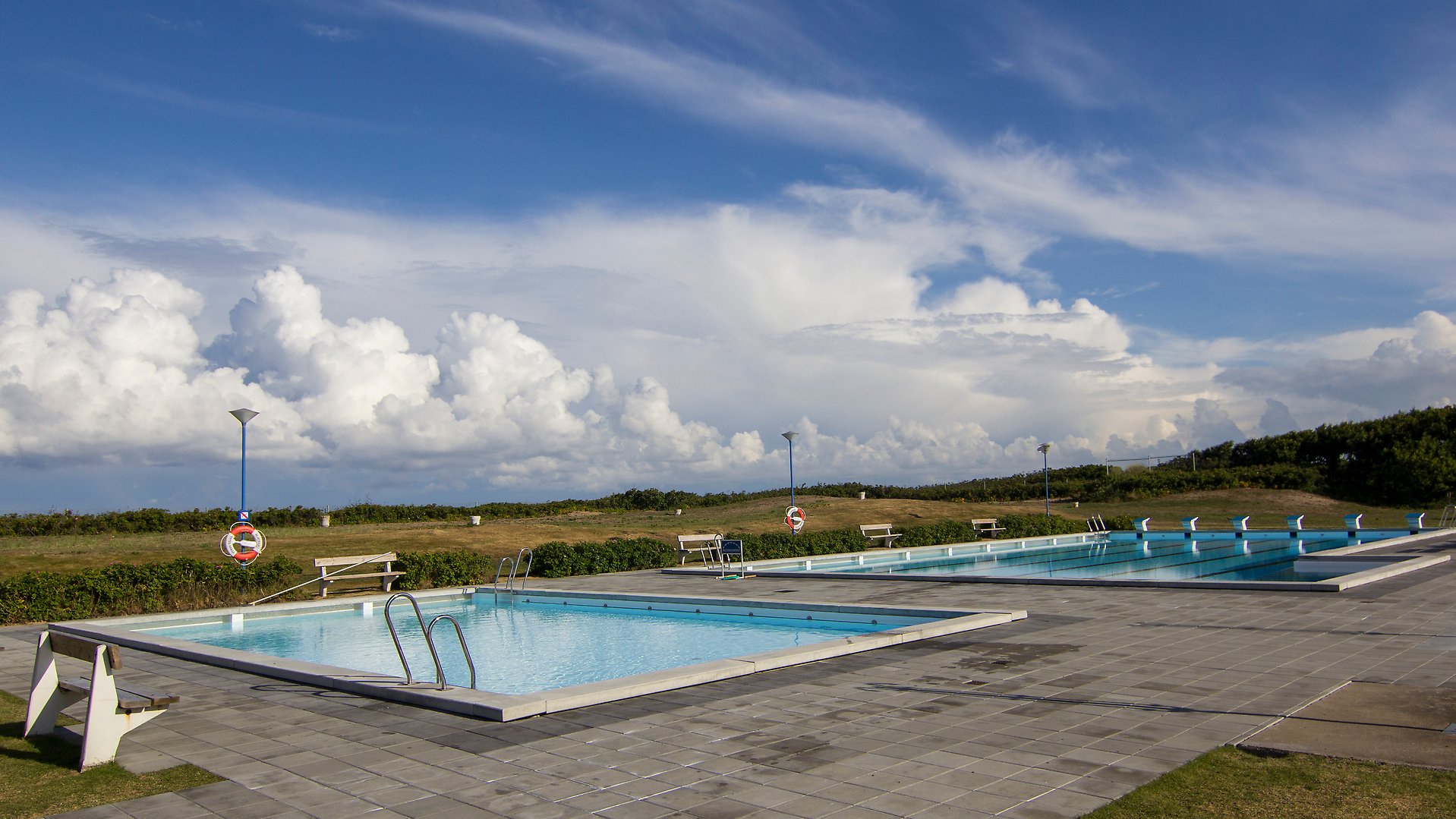 Bild över Vejbybadets mellan- respektive större bassäng. Blå himmel, delvis täckt av moln. Foto.