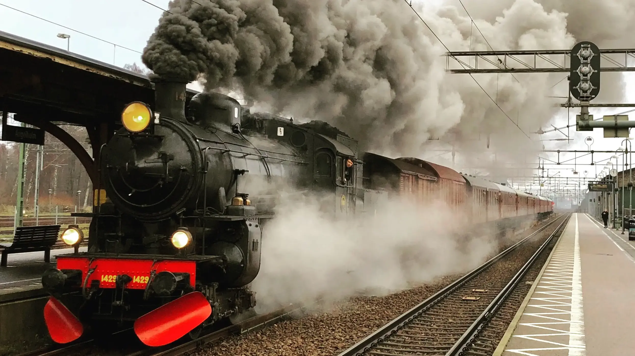 Ångtåg står på spåret vid en perrong och svart rök väller ut från taket på tåget. foto. 