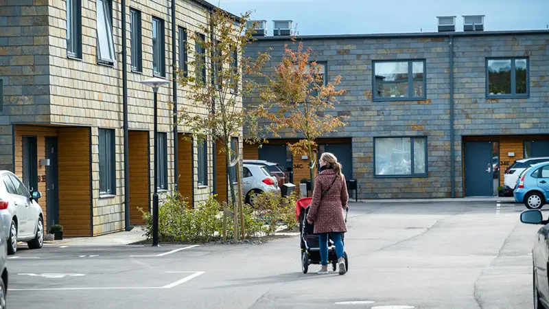 Kvinna med barnvagn går bland lägre nybyggda radhus. Ryggen emot. Foto. 
