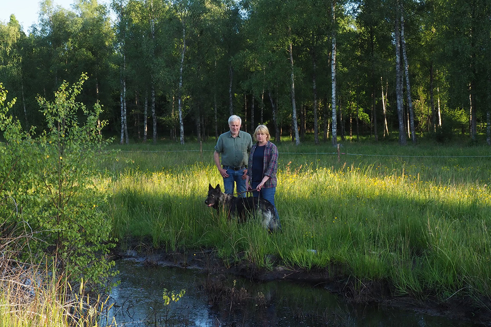 En man och kvinna i 50-60-årsåldern klädda i jeans och skjortor står i högt gräs med en schäferhund och tittar mot kameran. I förgrunden täcks marken av vatten. I bakgrunden syns ett ståltrådsstängsel och björkskog. Solen skiner och blå himmel skymtas. Foto.